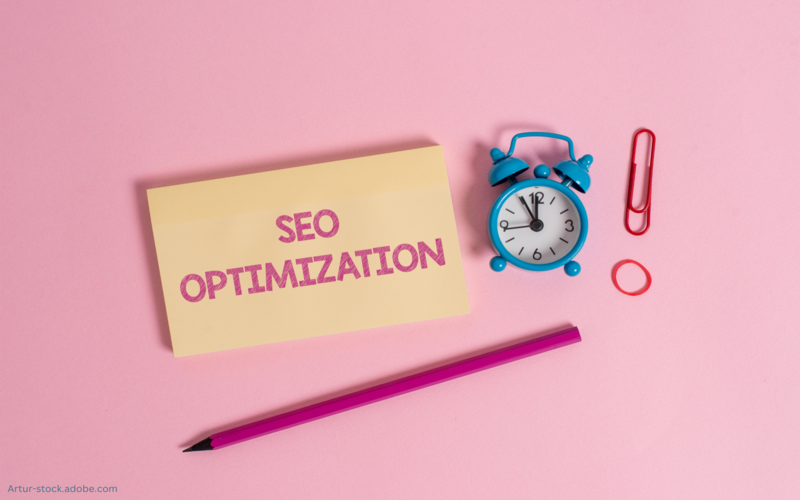 Auf dem Bild ist ein Zettel mit der Aufschrift "SEO Optimization" zu sehen. Daneben ist ein Wecker.